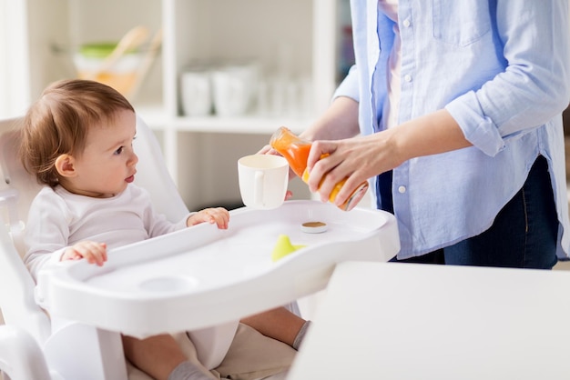 concetto di famiglia, cibo, alimentazione e persone - piccolo bambino e madre che versano il succo di carota naturale dalla bottiglia di vetro alla tazza nella cucina di casa