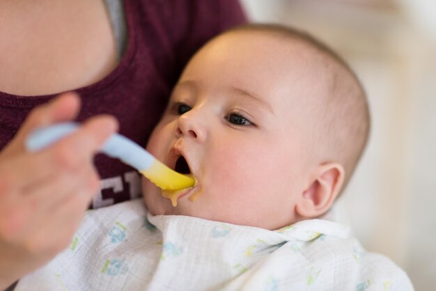 concetto di famiglia, cibo, bambino, alimentazione e genitorialità madre con cucchiaio che alimenta il piccolo bambino a casa