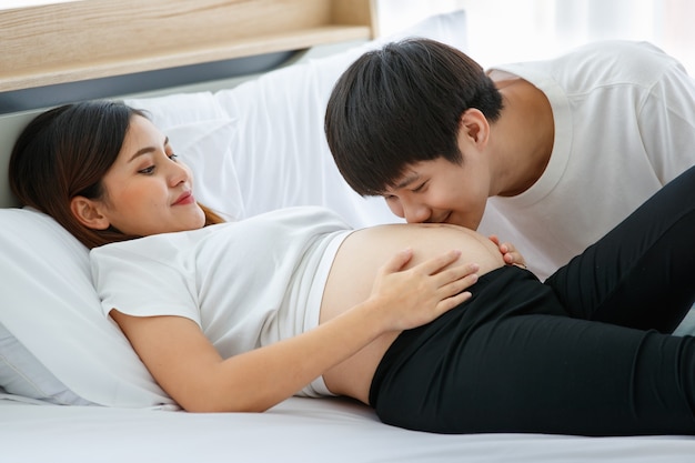 Concetto di famiglia di felicità. Immagine di una giovane coppia asiatica uomo e donna su un letto. Un bel marito che si bacia sulla pancia di una donna incinta mentre la sua bella moglie lo guarda con amore.