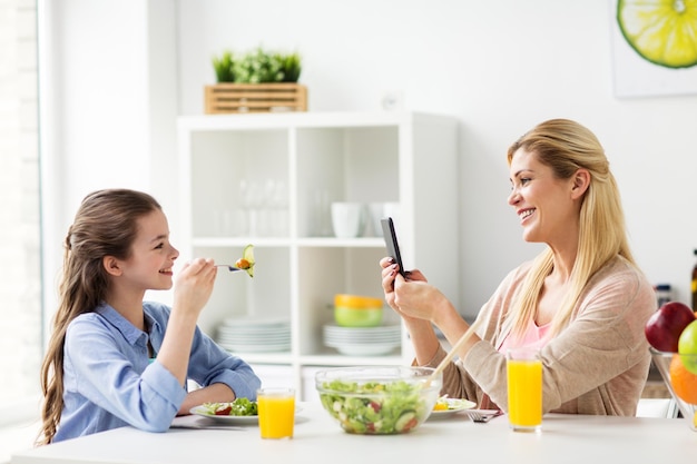 concetto di famiglia e persone - madre felice con smartphone che cena e fotografa sua figlia in cucina a casa