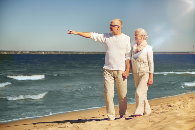 concetto di famiglia, età, viaggi, turismo e persone - coppia senior felice che cammina e punta il dito verso qualcosa sulla spiaggia estiva