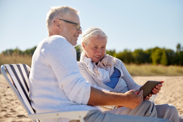 concetto di famiglia, età, viaggi, turismo e persone - coppia senior felice con computer tablet pc che riposa in sedie pieghevoli sulla spiaggia estiva