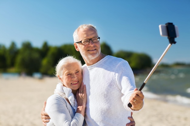 concetto di famiglia, età, viaggio, tecnologia e persone - felice coppia senior con smartphone selfie stick fotografare e abbracciare sulla spiaggia estiva