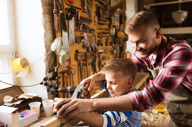 concetto di famiglia, falegnameria, falegnameria e persone - padre e figlio piccolo con aereo che lavora con tavola di legno in officina