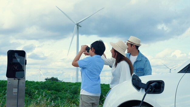 Concetto di famiglia felice e progressista che si gode il tempo al parco eolico con un veicolo elettrico Veicolo elettrico guidato da energia pulita e rinnovabile proveniente dal generatore eolico per la stazione di ricarica