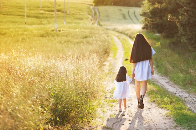 Concetto di famiglia felice. Mamma e bambino che tengono le mani e camminano la sera nei raggi del bel tramonto. Piccola figlia cammina con sua madre sulla strada. Campo giallo, sole al tramonto, giorno e sera d'estate