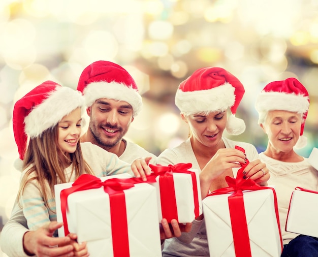 concetto di famiglia, felicità, generazione, vacanze e persone - famiglia felice in cappelli di Babbo Natale con scatole regalo seduto sul divano su sfondo di luci