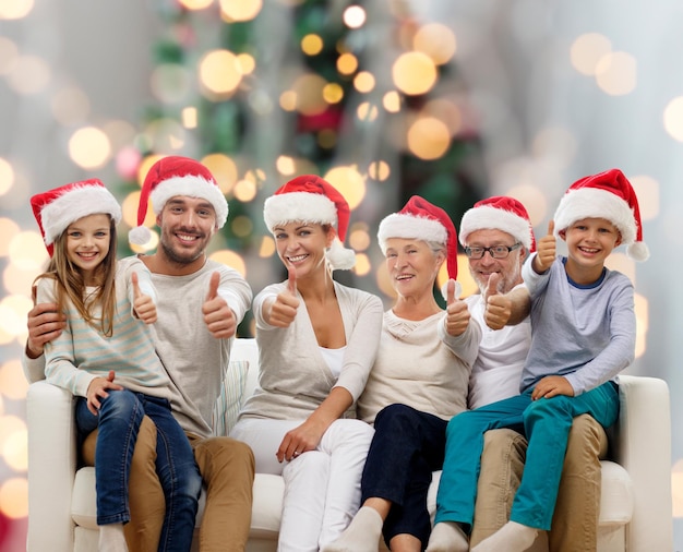concetto di famiglia, felicità, generazione, vacanze e persone - famiglia felice in cappelli di Babbo Natale seduto sul divano e mostrando il pollice in alto gesto su sfondo di luci ad albero