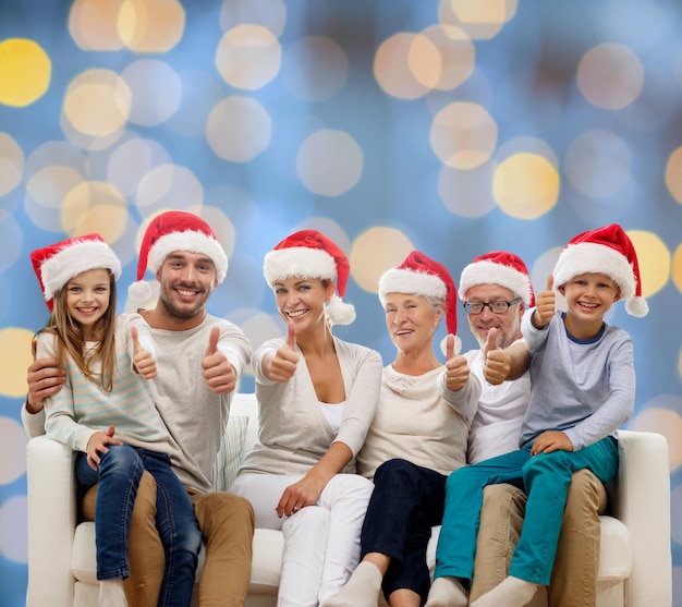 concetto di famiglia, felicità, generazione, vacanze e persone - famiglia felice in cappelli di Babbo Natale seduto sul divano e mostrando il pollice in alto gesto su sfondo di luci blu