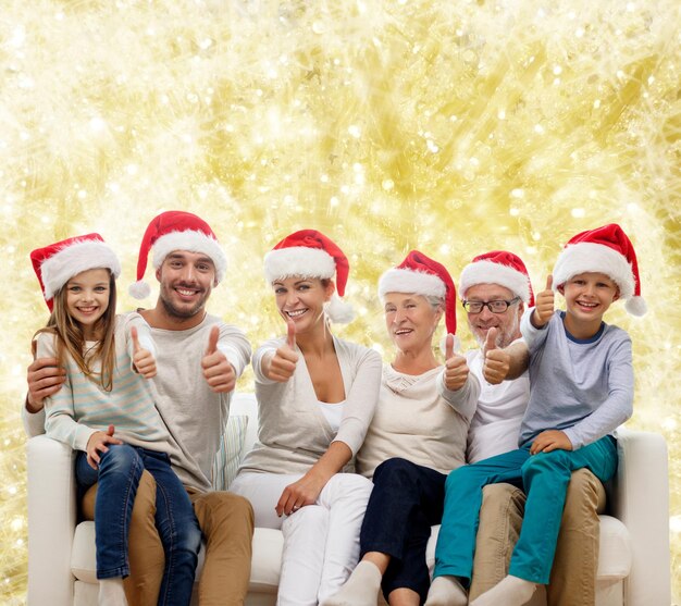 concetto di famiglia, felicità, generazione, vacanze e persone - famiglia felice in cappelli di Babbo Natale seduto sul divano e mostrando il pollice in alto gesto su sfondo di luci gialle