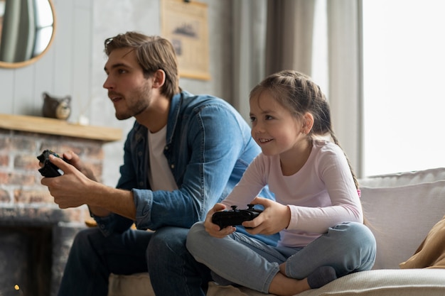 Concetto di famiglia, gioco e intrattenimento: padre felice e figlia piccola con gamepad che giocano a videogiochi a casa.