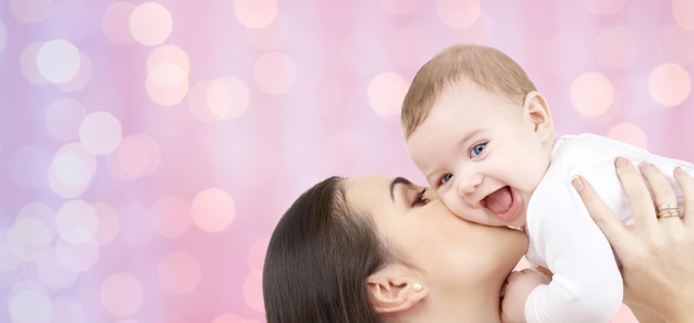 concetto di famiglia, maternità, bambini, genitorialità e persone - madre felice che bacia il suo bambino su sfondo di luci di vacanze rosa