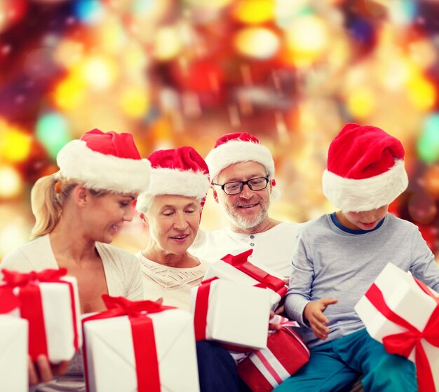 concetto di famiglia, natale, generazione, vacanze e persone - famiglia felice in cappelli di Babbo Natale con scatole regalo seduto su sfondo a luci rosse