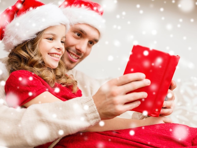 concetto di famiglia, natale, persone e vacanze - padre e ragazza sorridenti in cappelli di Babbo Natale che leggono un libro o la Bibbia a casa