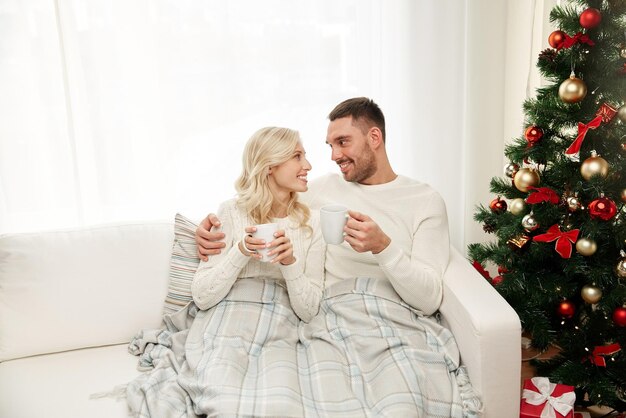 concetto di famiglia, natale, vacanze, amore e persone - coppia felice coperta di plaid che beve tè e si siede sul divano a casa