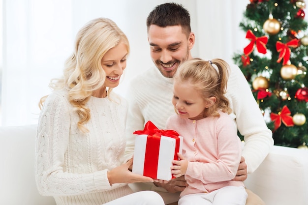 concetto di famiglia, natale, vacanze e persone - madre felice, padre e figlia piccola con confezione regalo seduta sul divano di casa