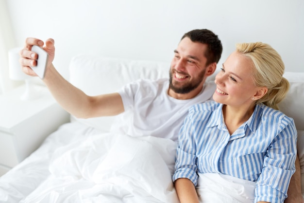 concetto di famiglia, tecnologia e persone - coppia felice con lo smartphone che si fa selfie a letto a casa