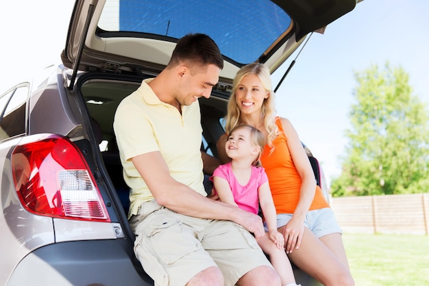 concetto di famiglia, trasporto, tempo libero, viaggio su strada e persone - uomo felice, donna e bambina seduti sul bagagliaio di un'auto berlina e parlare all'aperto