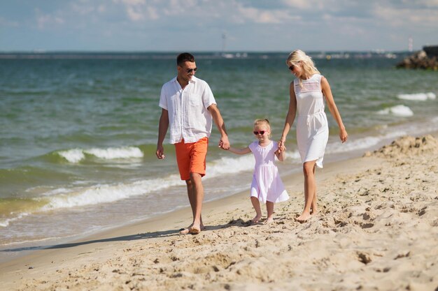 concetto di famiglia, vacanza, adozione e persone - uomo felice, donna e bambina in occhiali da sole che camminano sulla spiaggia estiva