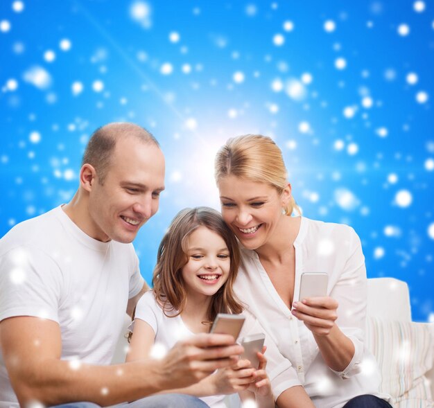 concetto di famiglia, vacanze di Natale, tecnologia e persone - madre, padre e bambina sorridenti con smartphone su sfondo blu innevato