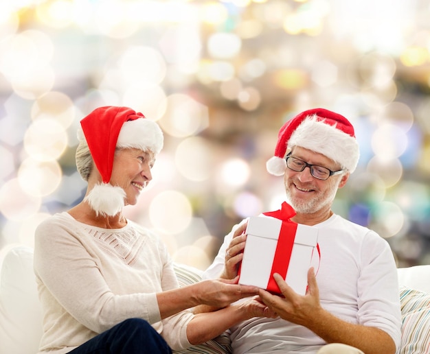 concetto di famiglia, vacanze, natale, età e persone - coppia senior felice in cappelli di Babbo Natale con confezione regalo su sfondo di luci