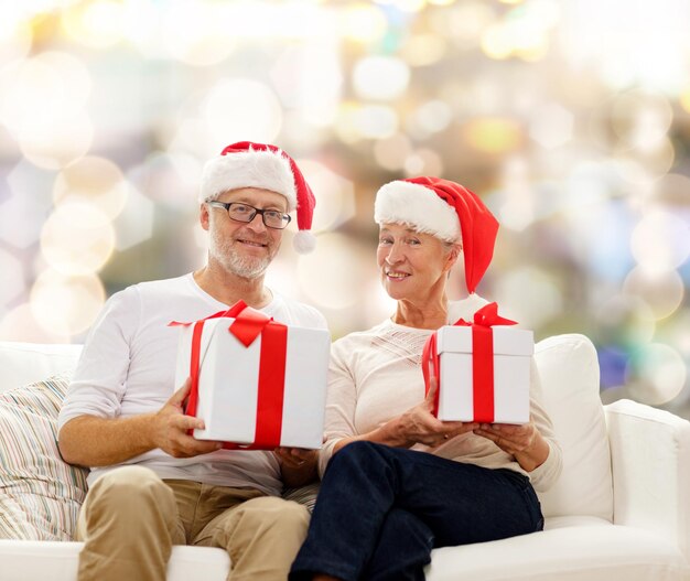 concetto di famiglia, vacanze, natale, età e persone - coppia senior felice in cappelli di Babbo Natale con scatole regalo su sfondo di luci