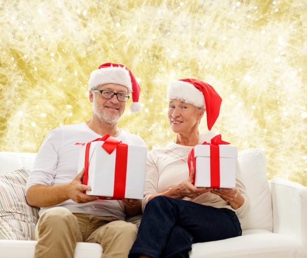 concetto di famiglia, vacanze, natale, età e persone - coppia senior felice in cappelli di Babbo Natale con scatole regalo su sfondo giallo