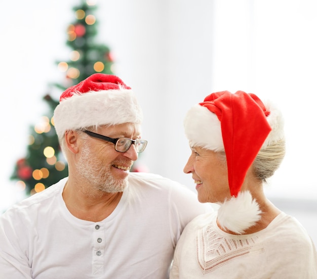 concetto di famiglia, vacanze, natale, età e persone - coppia senior felice in cappelli di Babbo Natale seduto sul divano sopra il soggiorno e lo sfondo dell'albero di natale