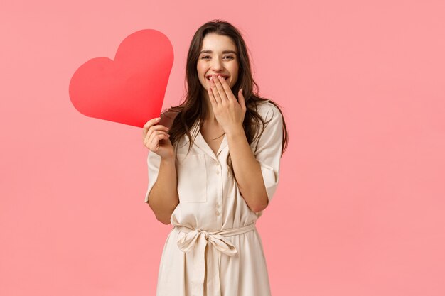 Concetto di felicità, relazione e romanticismo. Seducente donna abbastanza femminile con i capelli ricci scuri, indossa un abito, ridendo la bocca della copertina come una risatina, con in mano un simpatico cuore rosso in cartone, parete rosa