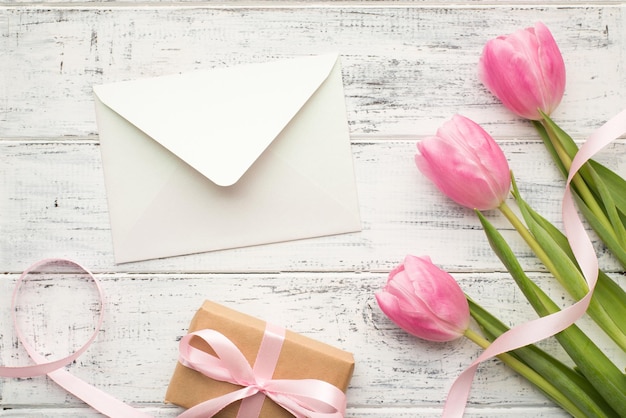 Concetto di festa della mamma felice. Laici piatta aerea vista ravvicinata foto di tre fiori di colore pastello con nastro kraft avvolto presente sulky e busta bianca aperta sul tavolo di legno