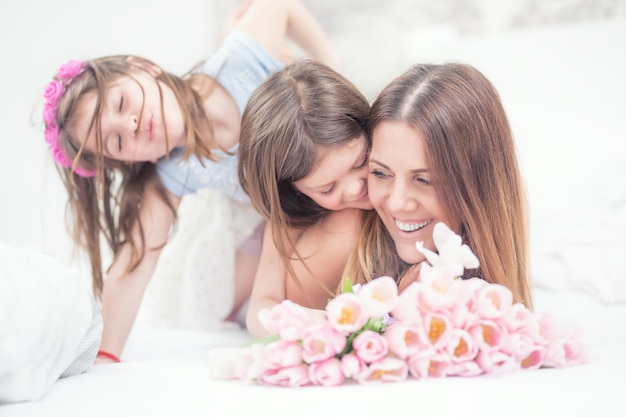 Concetto di festa della mamma felice. Mamma con due simpatiche figlie gemelle sul letto in camera da letto e un mazzo di tulipani di fiori.