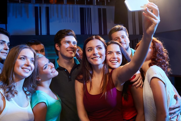 concetto di festa, tecnologia, vita notturna e persone - amici sorridenti con smartphone che prendono selfie nel club