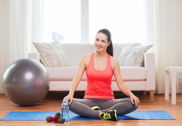 concetto di fitness, casa e dieta - sorridente ragazza seduta sul tappetino con attrezzature sportive a casa