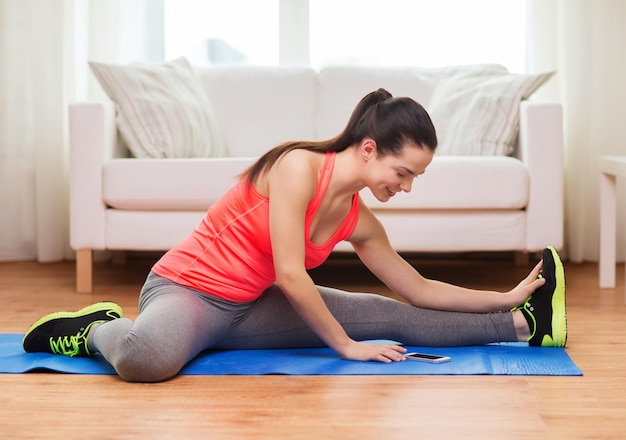 concetto di fitness, casa, tecnologia e dieta - ragazza sorridente che si allunga sul pavimento con lo smartphone a casa