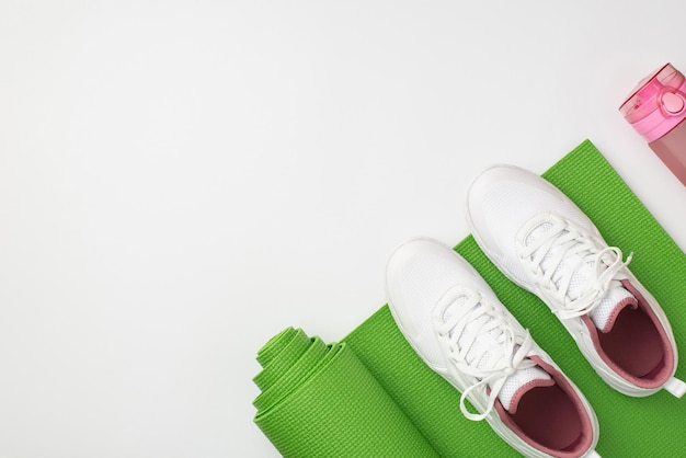 Concetto di fitness Foto vista dall'alto di scarpe sportive bianche bottiglia rosa e tappetino verde per esercizi su sfondo bianco isolato con copyspace