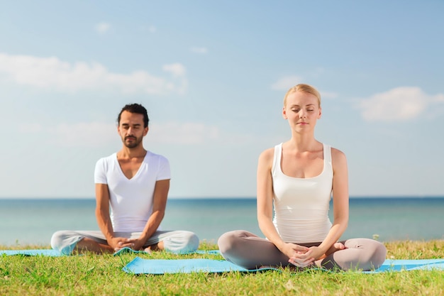 concetto di fitness, sport, amicizia e stile di vita - coppia sorridente che fa esercizi di yoga seduti su stuoie all'aperto