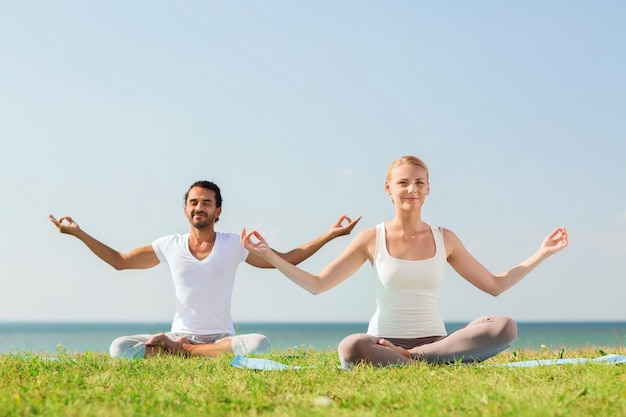 concetto di fitness, sport, amicizia e stile di vita - coppia sorridente che fa esercizi di yoga seduti su stuoie all'aperto