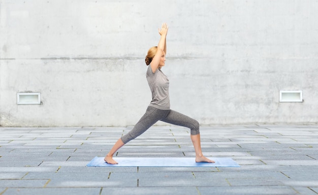 concetto di fitness, sport, persone e stile di vita sano - donna che fa la posa del guerriero yoga sul tappetino su sfondo di strada urbana