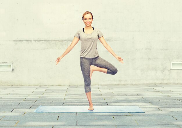 concetto di fitness, sport, persone e stile di vita sano - donna che fa yoga nella posa dell'albero sul tappetino su sfondo di strada urbana