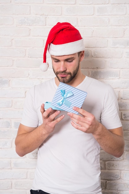 Concetto di giorno di Santo Stefano. Scatola di Natale aperta uomo con nastro azzurro. Fare regali, auguri per le vacanze. Macho in cappello della santa con il presente sul muro di mattoni bianco. Natale, preparazione e celebrazione del nuovo anno.