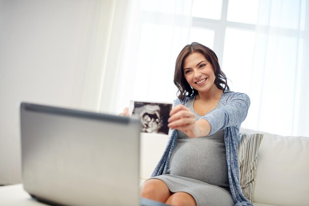 concetto di gravidanza, maternità, persone e medicina - donna incinta felice con computer portatile che ha una videochiamata e mostra l'immagine ecografica a casa