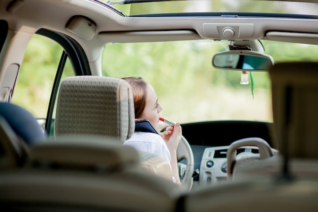 Concetto di guida pericolosa. Adolescente dai capelli rossi del driver della giovane donna che dipinge le sue labbra che fanno stendere il trucco mentre guidando l'automobile