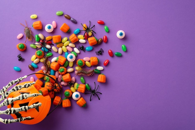 Concetto di Halloween Foto vista dall'alto della mano scheletro che tiene cesto di zucca con occhi di caramelle millepiedi scarafaggio e ragni su sfondo viola isolato