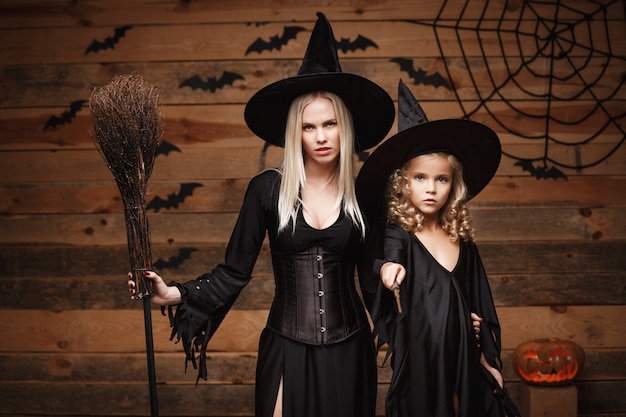 Concetto di Halloween - madre allegra e sua figlia in costumi della strega che celebrano posa di Halloween con zucche curve sopra pipistrelli e ragnatela sullo sfondo di legno di studio.