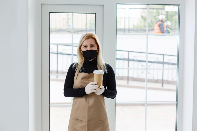 Concetto di imprenditore - Il bellissimo barista caucasico in maschera facciale offre caffè caldo nella moderna caffetteria