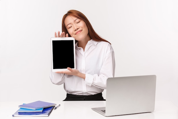 Concetto di imprenditori di carriera, lavoro e donne. Il ritratto del primo piano della donna asiatica sorridente vaga si siede alla scrivania, chiude gli occhi sognando di qualcosa, abbracciando la compressa con la foto sul display