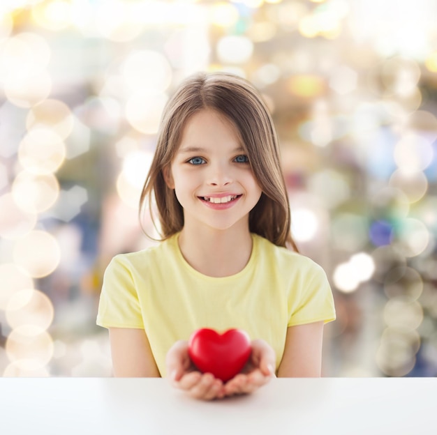 concetto di infanzia, amore, carità, vacanze e persone - bambina sorridente che si siede e tiene il cuore rosso su sfondo scintillante