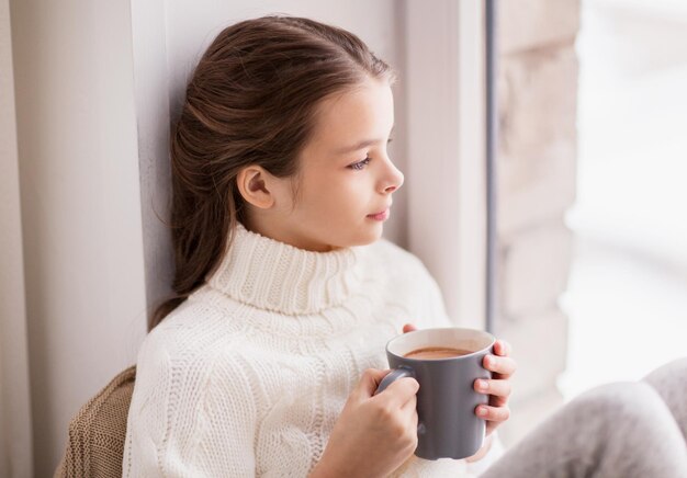 concetto di infanzia e persone - bella ragazza con tazza di cacao in maglione seduto alla finestra di casa in inverno