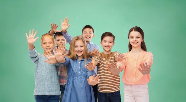 concetto di infanzia, educazione, amicizia, gesto e persone - bambini sorridenti felici che agitano le mani sopra il fondo verde del bordo di gesso della scuola