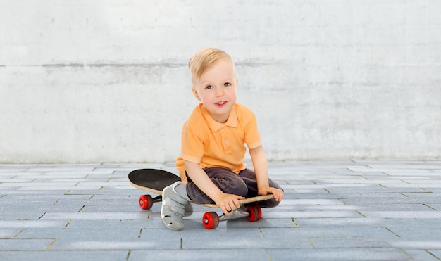 concetto di infanzia, sport, tempo libero e persone - ragazzino felice seduto su skateboard su sfondo di strada cittadina